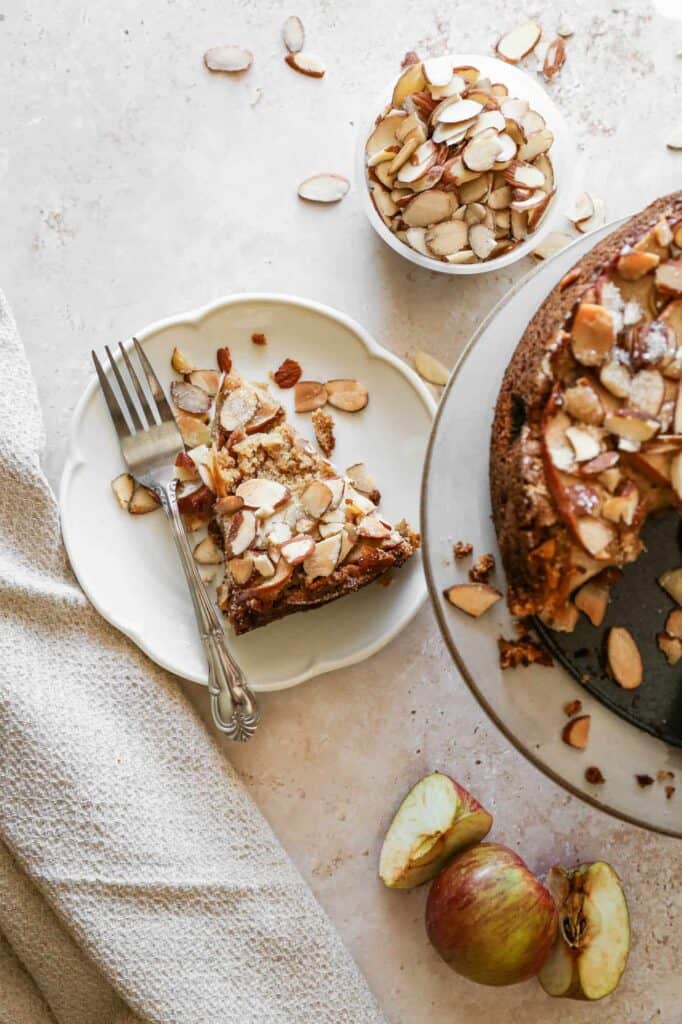 a slice of gluten free apple almond cake next to the cake and a bowl of almond slivers and apple slices