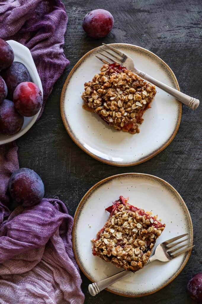 Two plates of gluten free plum cake with a bowl of fresh plums