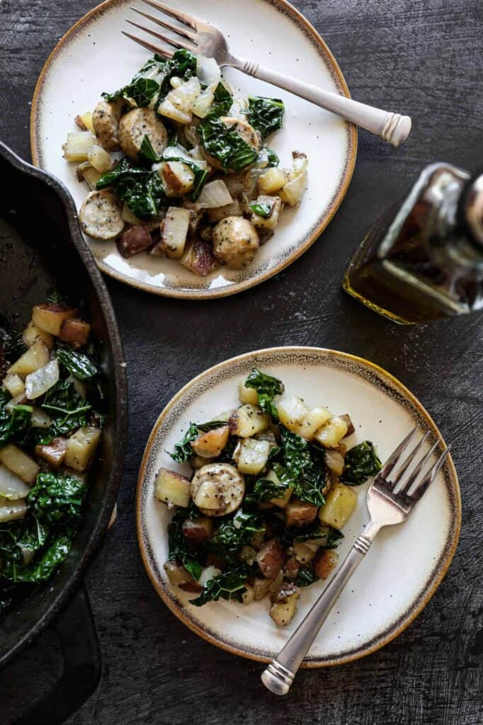 two plates of gluten free sausage, kale and potato skillet with a small bottle of olive oil