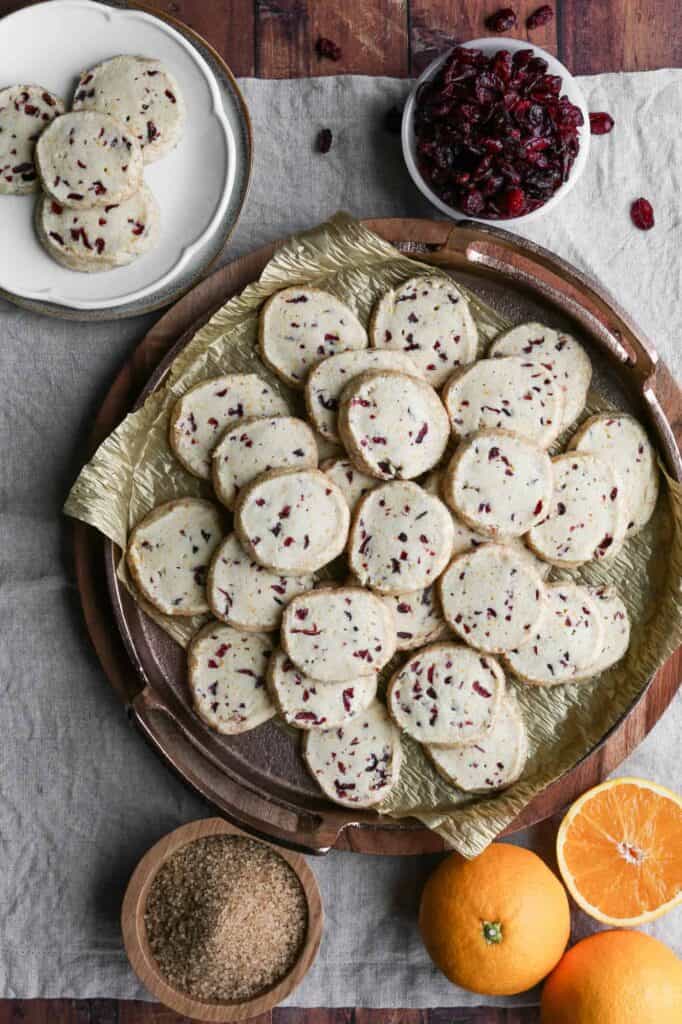 a tray of gluten free orange cranberry butter cookies with a small plate of cookies, a bowl of dried cranberries and three oranges