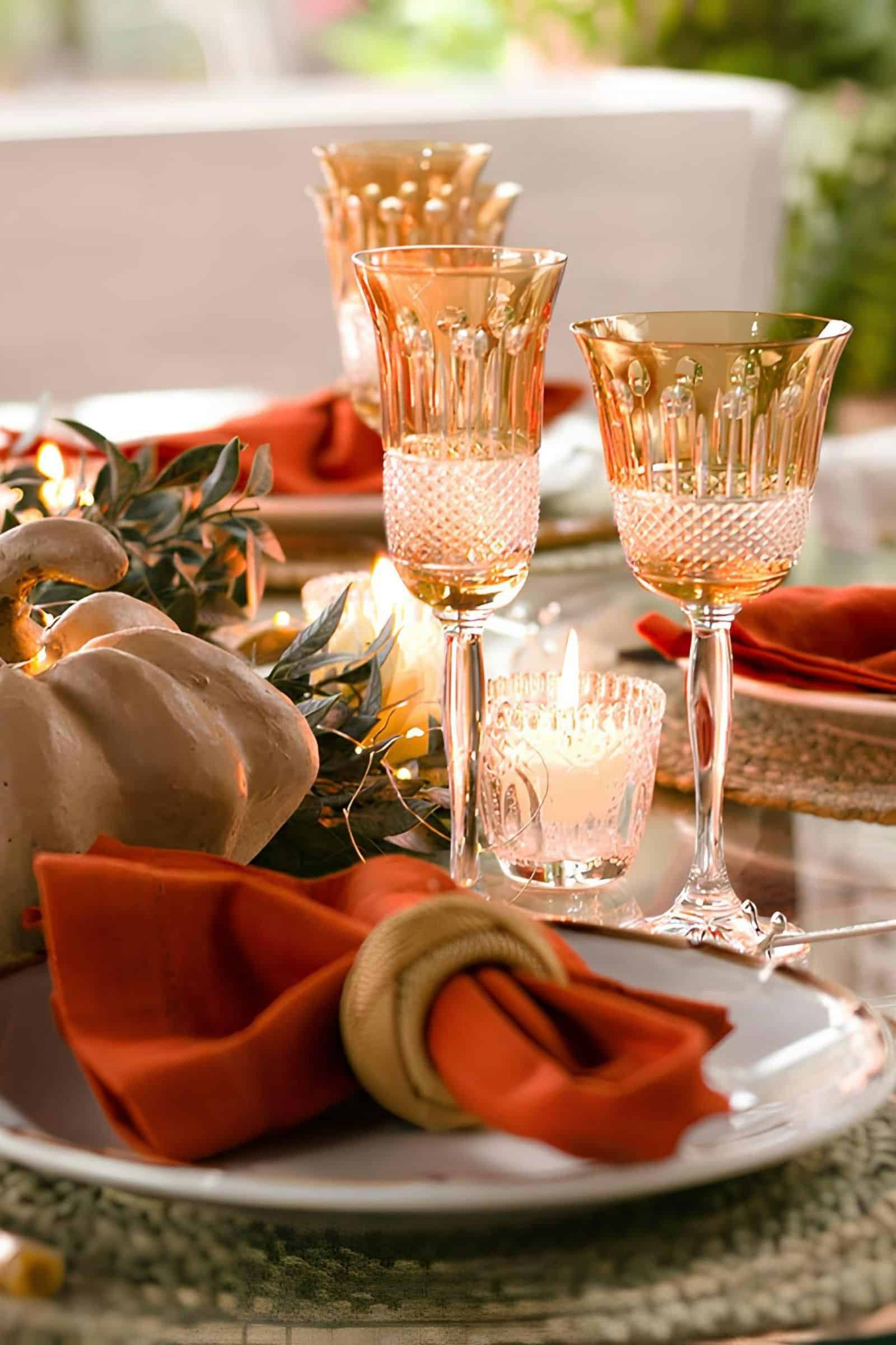 a table set with plates, orange napkins, pumpkins and amber stemware with tea light candles in cut glass holders