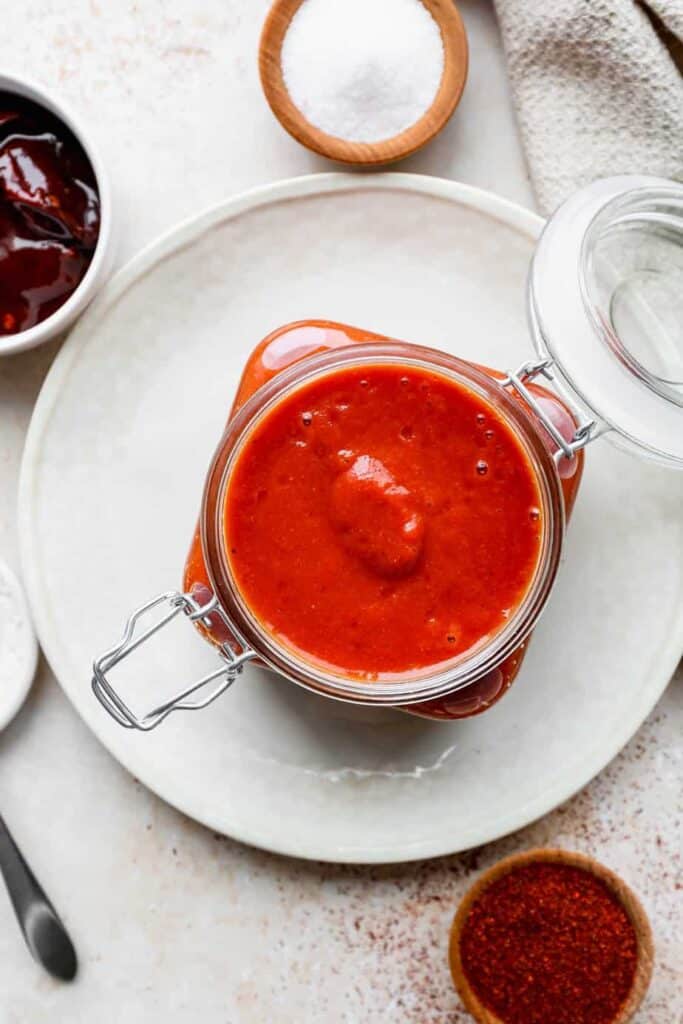 a glass jar of bright red gluten free homemade enchilada sauce with little dishes of salt, poblano peppers, pepper and chili powder around it