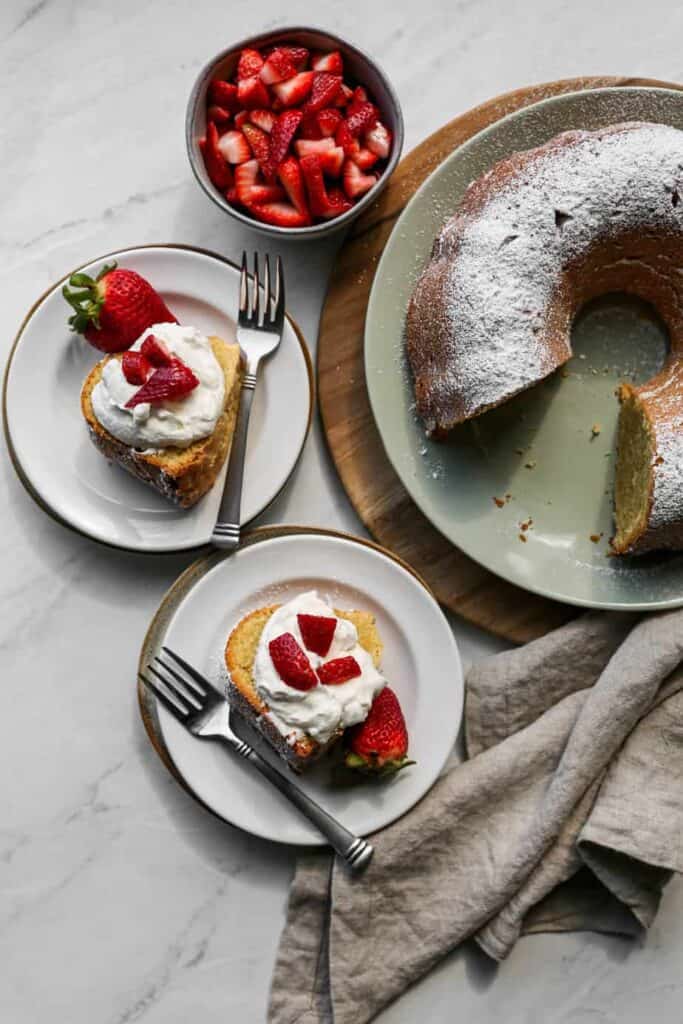 a gluten free cream cheese pound cake on a light blue platter with two slices on white plates with sliced strawberries and homemade whipped cream