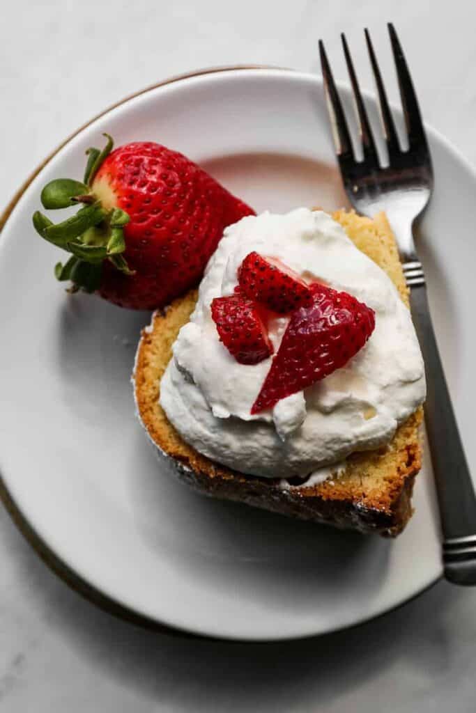 a slice of gluten free cream cheese pound cake on a white plate with sliced strawberries and homemade whipped cream on top