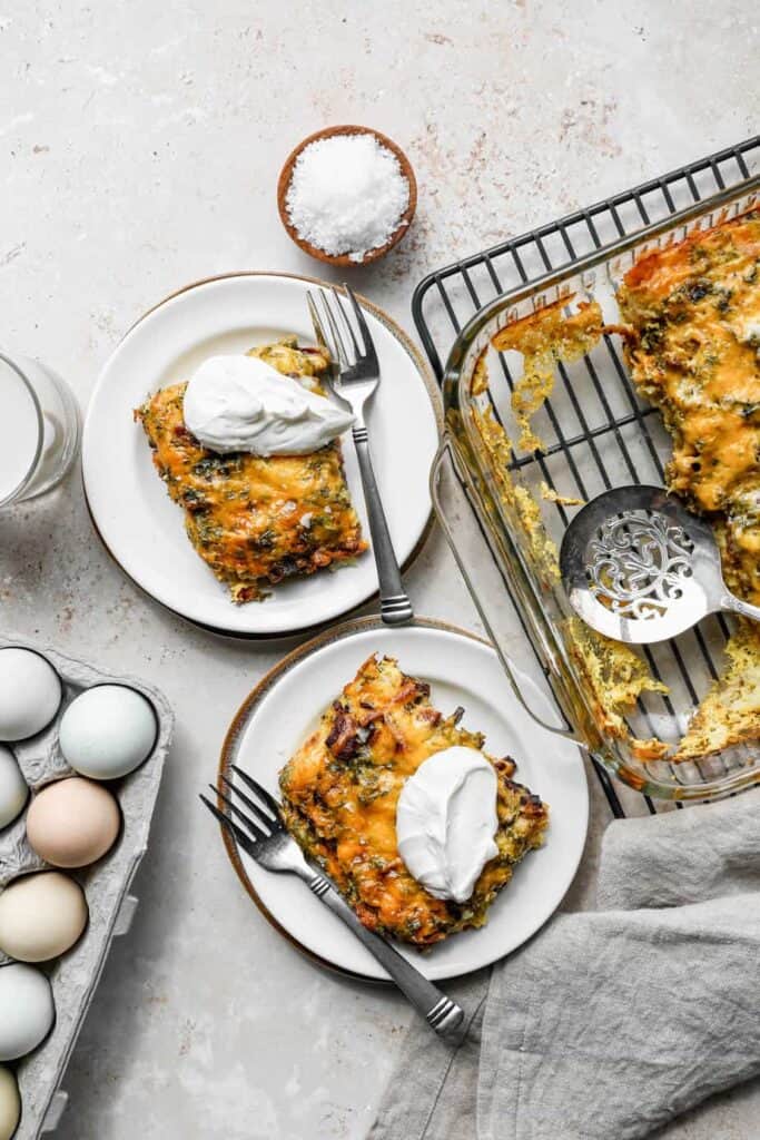 two white plates with slices of gluten free tater tot breakfast casserole with dollops of sour cream on top next to a 9x13 baking dish of the casserole