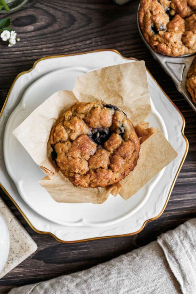 a white plate with a gluten free blueberry oat muffin next to a tin of muffins