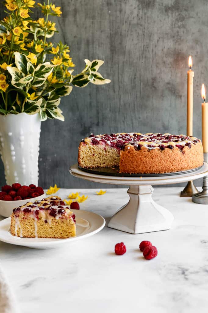 a gluten free raspberry cornmeal cake on a cake stand with a slice on a white plate with a bouquet of yellow flowers next to it