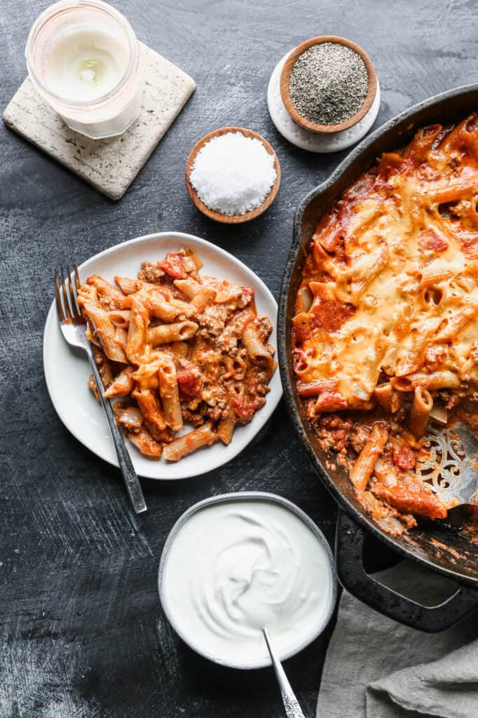 a cast iron skillet and a small white plate of gluten free taco pasta bake with a pinch bowl of salt and pepper and a bowl of sour cream