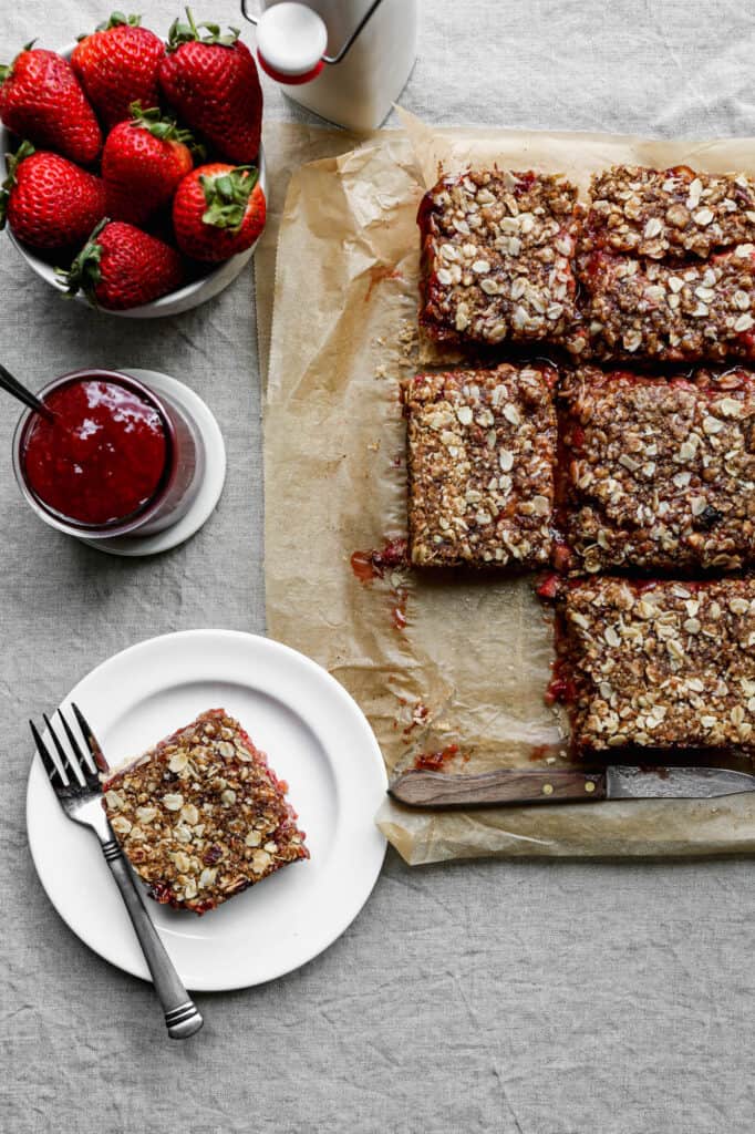 gluten free strawberry jam bars cut into squares on parchment paper with a bowl of fresh strawberries, a jar of strawberry jam and a bottle of milk