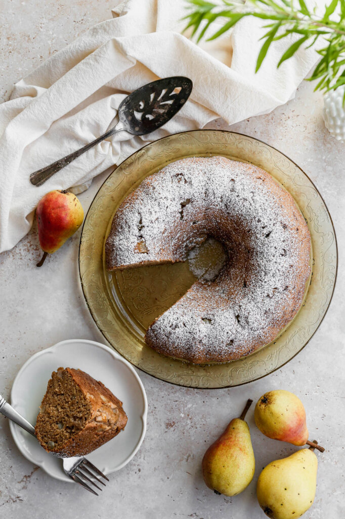 a gluten free pear bundt cake on a yellow carved glass plate with powdered sugar sprinkled on top next to a few fresh pears