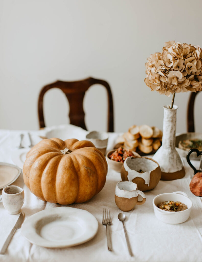 a table set for Thanksgiving dinner with a large orange pumpkin and vase of dried hydrangea