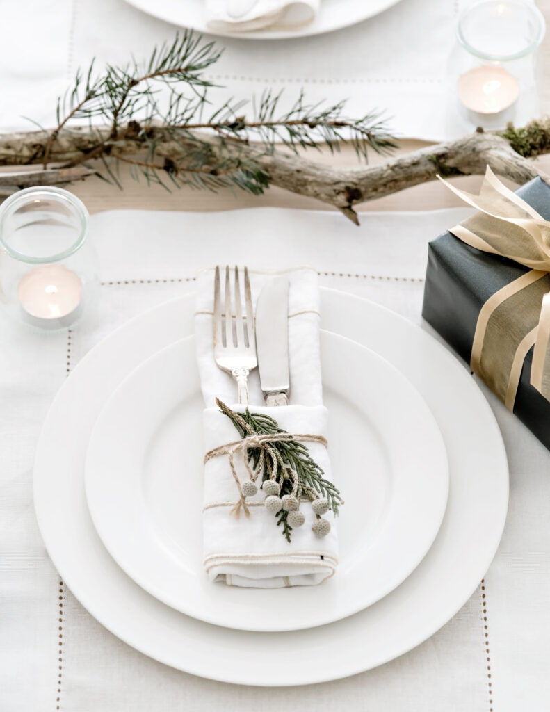 two white plates with a bundle of silver ware with a white napkin, twine and a pine cone next to white candles and a stick with an evergreen branch attached to it and a gift wrapped in green wrapping paper with a gold bow