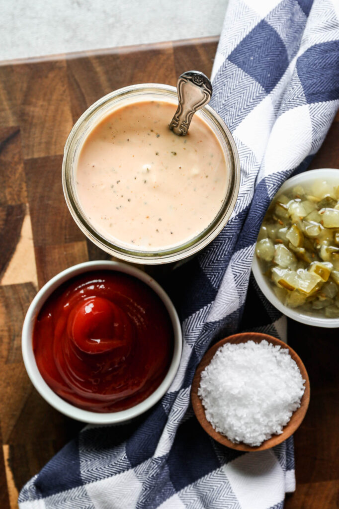 a jar of gluten free burger sauce next to a bowl of dill relish, a bowl of ketchup and dish of salt