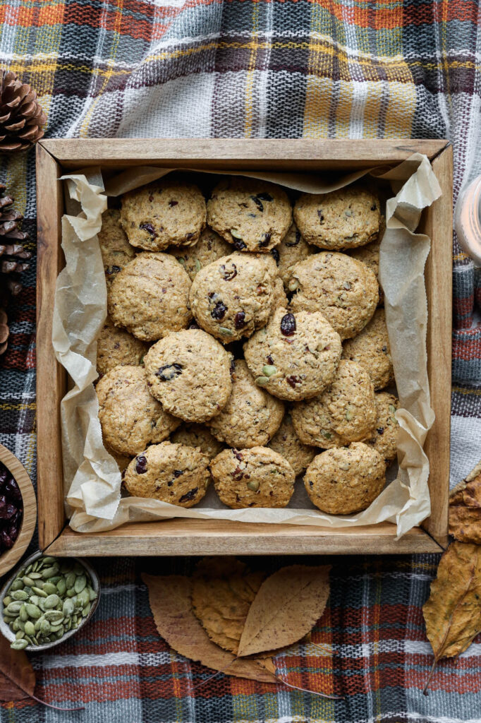 a wooden box lined with parchment paper of gluten free pumpkin breakfast cookies on a plaid blanket next to a bowl of pipits and a bowl of cranberries