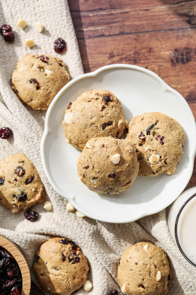 a white plate with three gluten free white chocolate cranberry cookies surrounded by other cookies and white chocolate chips and dried cranberries scattered around them