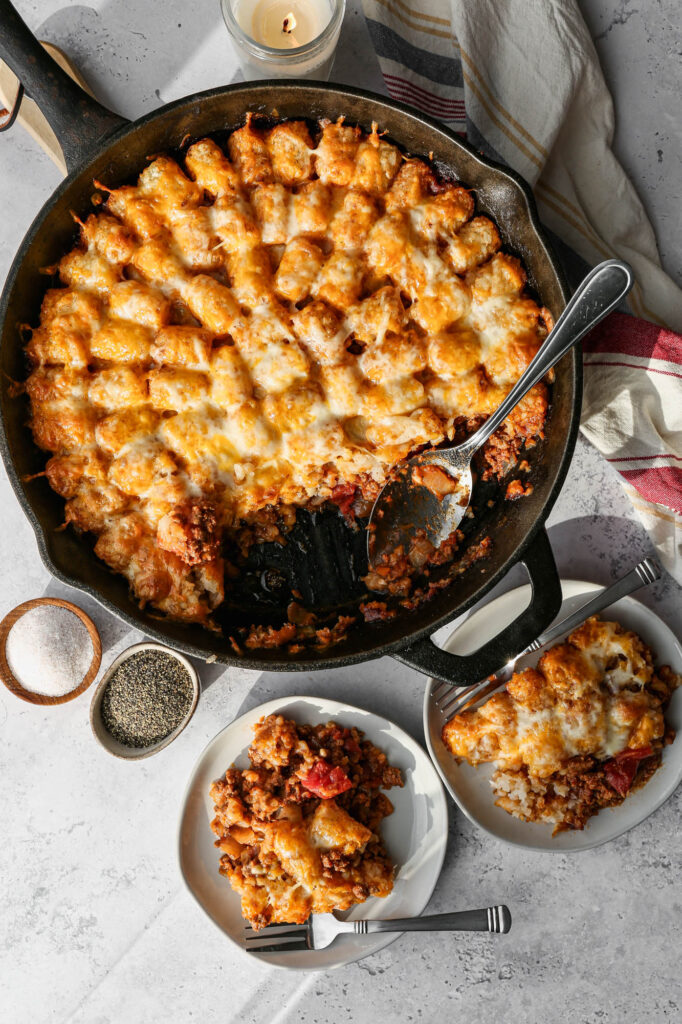 a cast iron skillet of gluten free cheesy tater tot casserole next to two white plates of casserole
