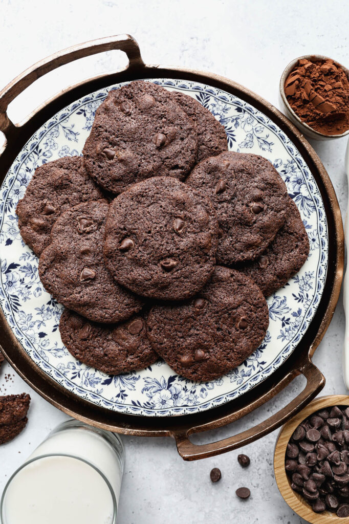a blue floral plate of gluten free double chocolate cookies next to a small bowl of cocoa powder, a glass of milk and a small wooden bowl of chocolate chips