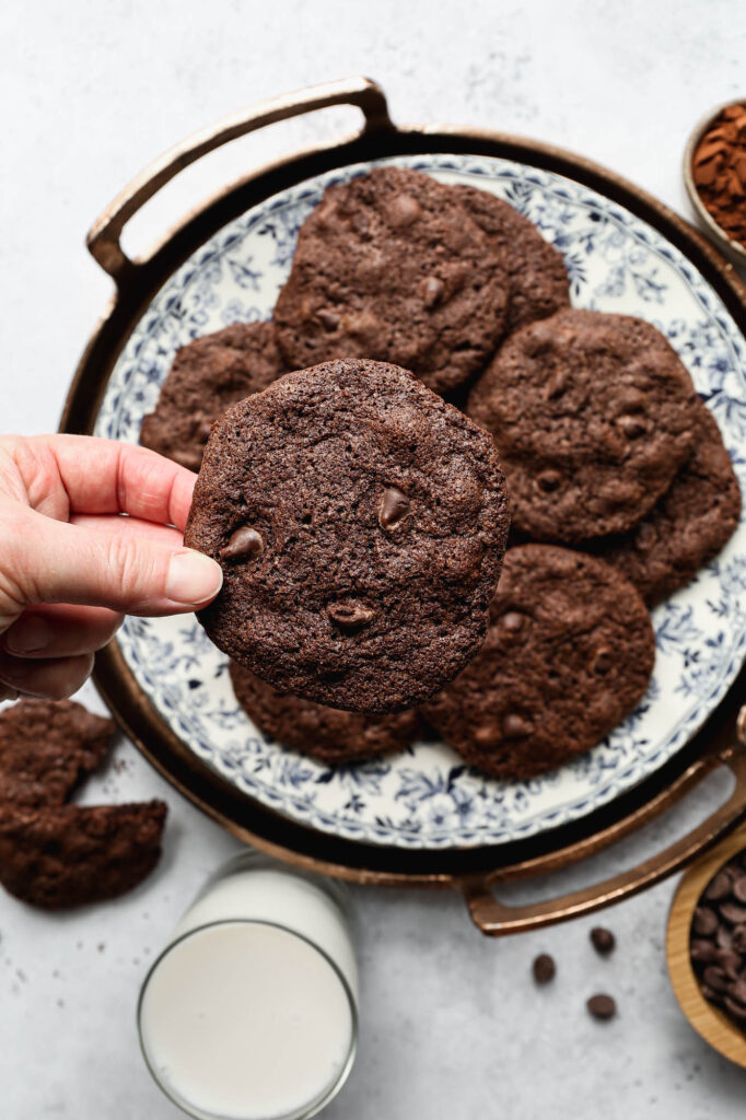 a blue floral plate of gluten free double chocolate cookies next to a small bowl of cocoa powder, a glass of milk and a small wooden bowl of chocolate chips