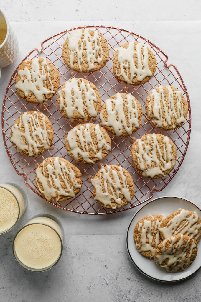 a copper cooling rack of gluten free eggnog cookies with eggnog icing drizzled overtop next to a white plate of cookies