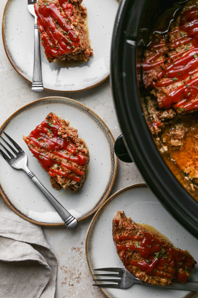 gluten free slow cooker meatloaf in a black crockpot with three white plates of meatloaf next to it