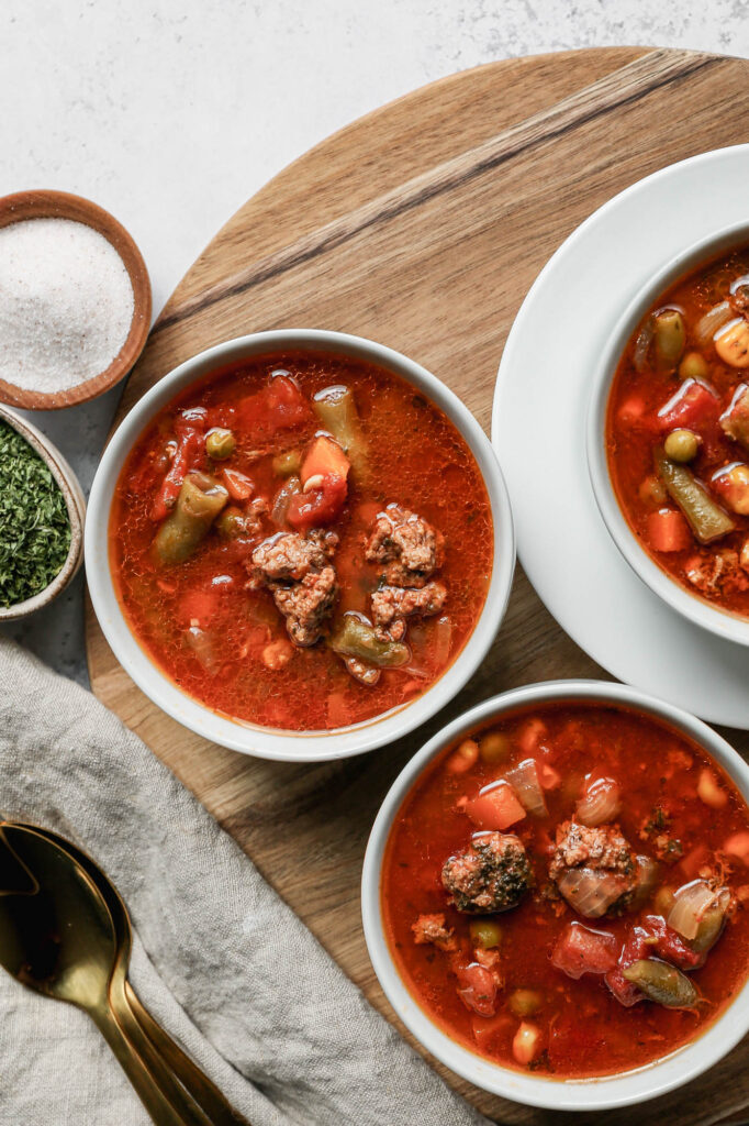 three white bowls of gluten free instant pot beef and vegetable soup sitting on a round wooden cutting board next to yellow onions