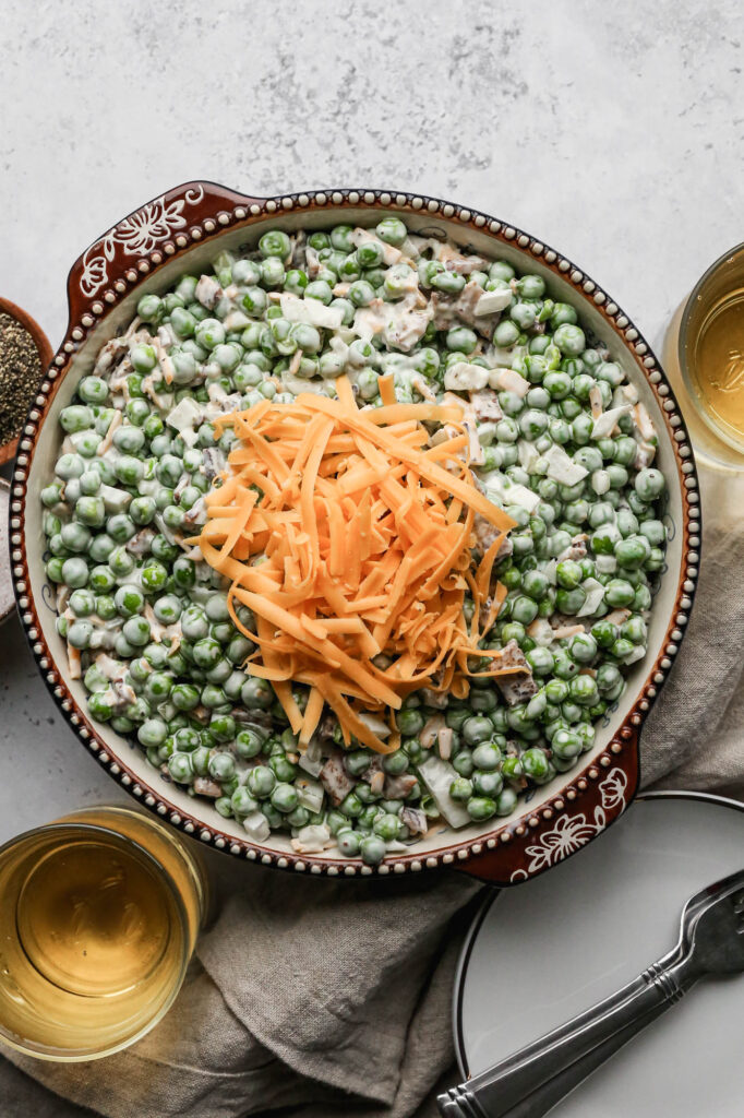 a bowl of easy pea salad with shredded cheddar cheese sprinkled on top next to a little wooden bowl of pepper and a little ceramic bowl of salt