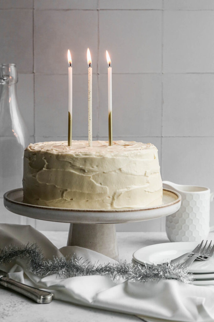 a gluten free eggnog cake with white chocolate frosting on a cream cake stand with white plates next to it and three white and gold candles lit on top