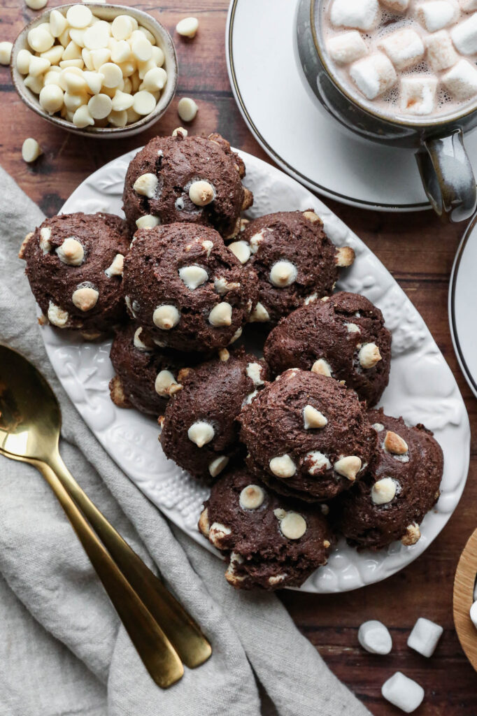 a small white platter of gluten free hot cocoa cookies next to two mugs of hot cocoa with small marshmallows on top, a little bowl of white chocolate ships and a wooden bowl of marshmallows