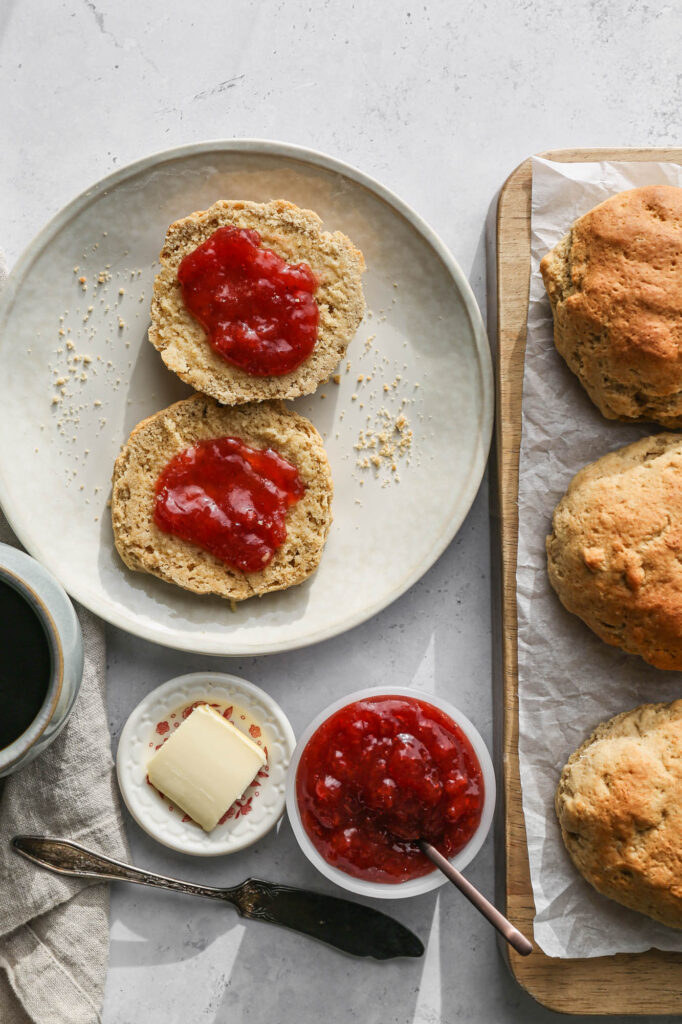 a cream plate with a gluten free millet drop biscuit cut in half with butter and strawberry jam on top next to a wood cutting board of biscuits and a small white bowl of strawberry jam