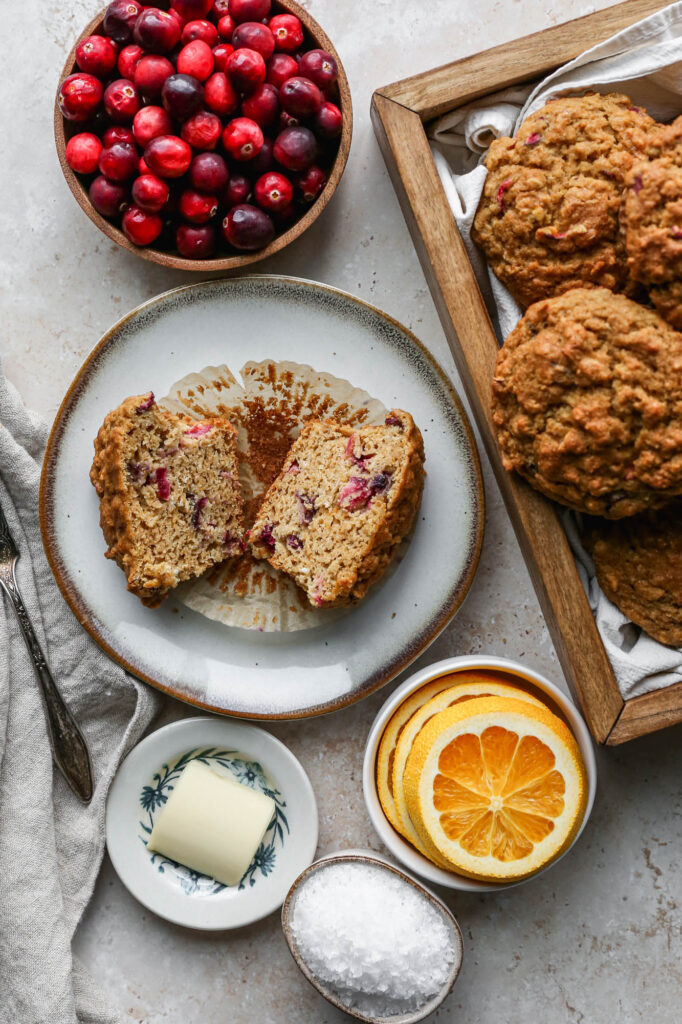 a small plate with a gluten free cranberry orange muffin cut in half next to a wooden box of muffins, a wooden bowl of cranberries, a little plate with a pat of butter and a small bowl of orange slices