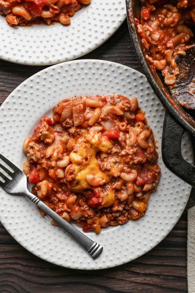 a cast iron skillet of gluten free sloppy joe casserole next to two white plates of casserole and a small bowl of salt and a small bowl of black pepper