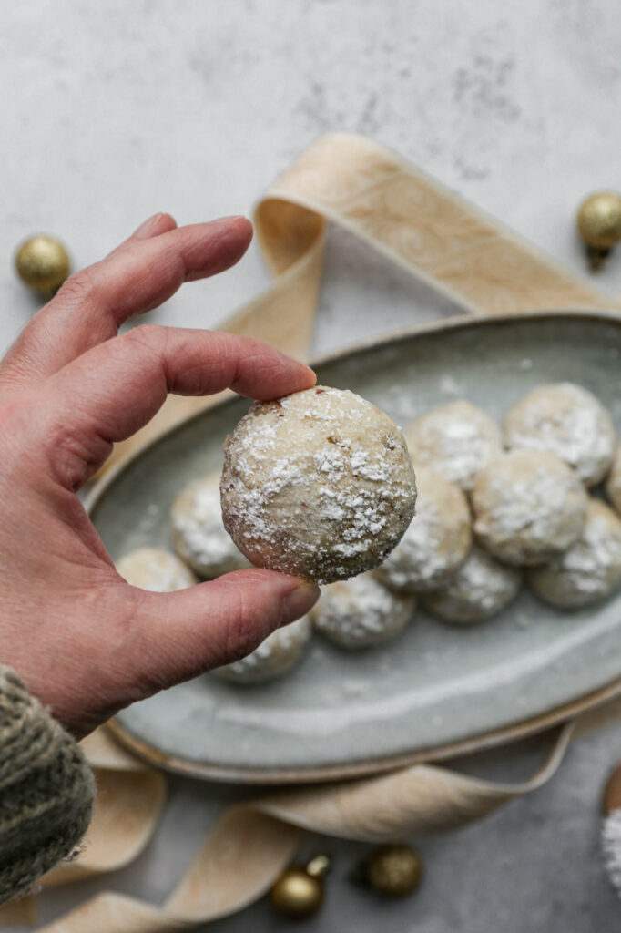 a platter of gluten free snowball cookies with cream patterned ribbon and small, round gold ornaments around it sitting next to a small white bowl of powdered sugar