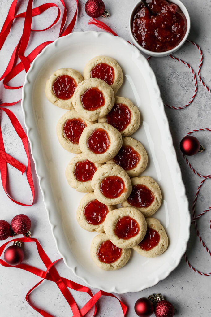 a white platter of gluten free thumbprint cookies with strawberry jam in the center next to red ribbon, a small white bowl of strawberry jam and some small red christmas ornaments