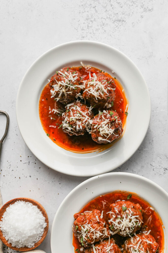 two white plates of gluten free slow cooker barbecue meatballs next to a white crockpot of meatballs and a cheese grater with parmesan cheese