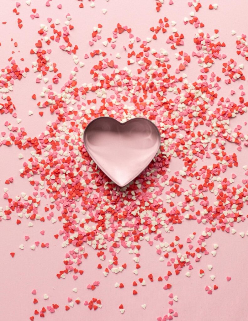 a pink background with a heart shaped cookie cutter and red, white and pink heart shaped sprinkles around it
