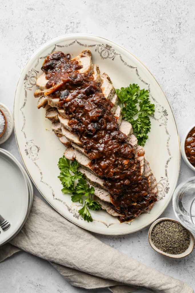 a cream platter of pork tenderloin with prune sauce next to a stack of plates and a small white bowl of sauce