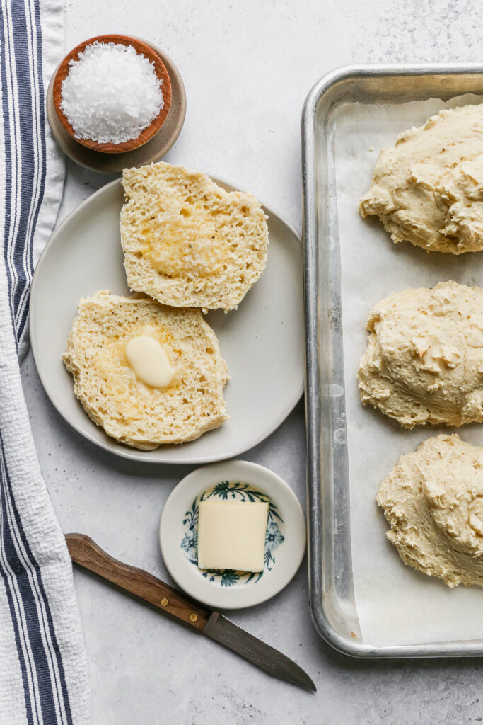 a white plate with a gluten free buttermilk drop biscuit cut in half with butter on each side next to a baking sheet of biscuits and a small wooden bowl of salt.