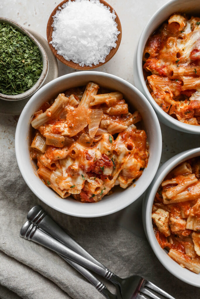 three white bowls of gluten free chicken parmesan pasta casserole next to a small bowl of salt and a small bowl of dried parsley.