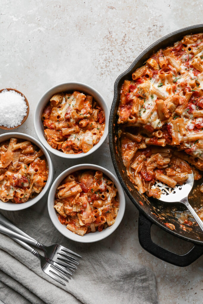 a cast iron skillet of gluten free chicken parmesan pasta casserole next to three white bowls of pasta.