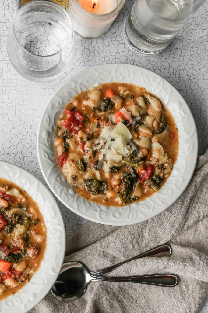 two white bowls of gluten free instant pot Italian style pasta and bean soup next to a glass of sparkling water and two soup spoons.