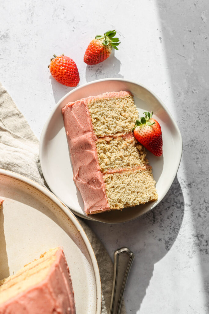 a white plate with a slice of gluten free vanilla cake with pink buttercream frosting next to fresh strawberries.