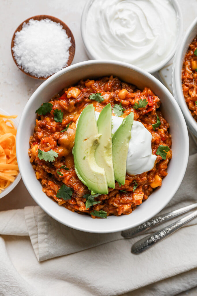 two white bowls of instant pot mexican style beef and rice next to a small white bowl of sour cream and a small white bowl of shredded cheddar cheese.