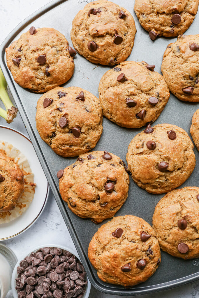 a muffin tin of gluten free chocolate chip banana muffins next to a small bowl of chocolate chips and white plate with a muffin on it.