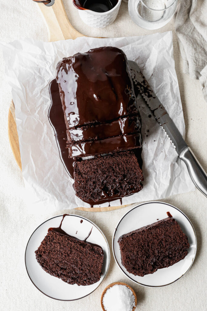 a gluten free chocolate pound cake with chocolate glaze poured over top next to two white plates with slices on them.
