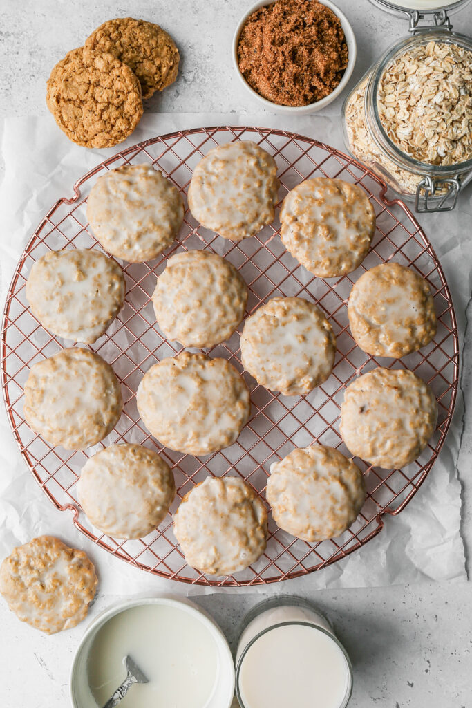 a round copper cooling rack with gluten free old fashioned iced oatmeal cookies next to a small white bowl of icing, a glass of milk, a small white bowl of brown sugar and a glas