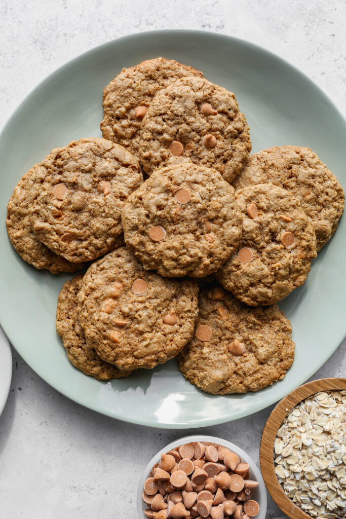 a sage green plate of gluten free scotchie cookies next to a small white bowl of butterscotch chips and a small wooden bowl of gluten free rolled oats.