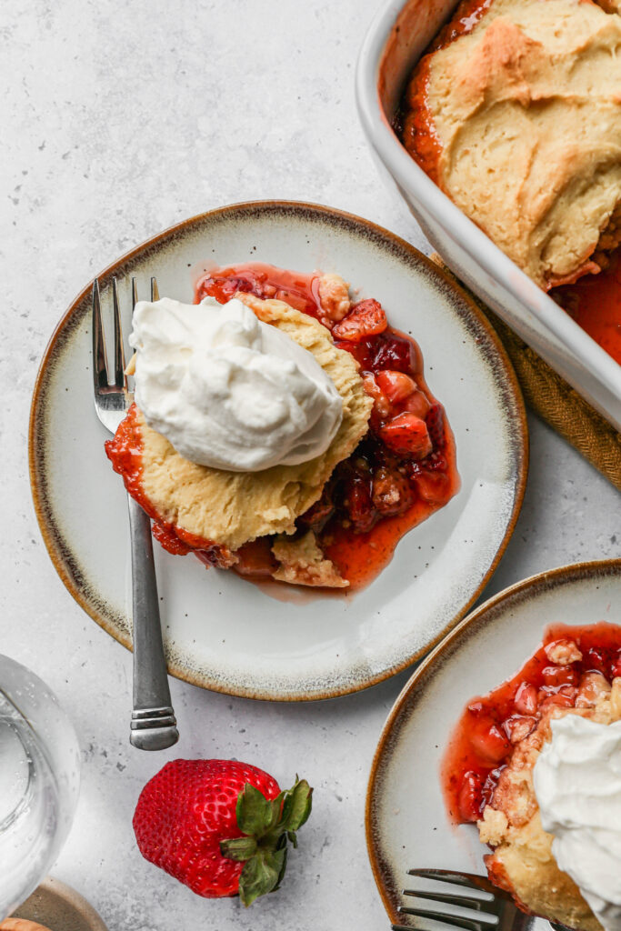 a plate of gluten free strawberry cobbler with homemade whipped cream on top next to a white baking dish of cobbler.