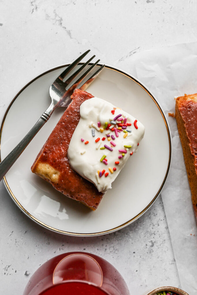 a slice of gluten free easter poke cake on a white plate next to a small bowl of bright colored sprinkles and a stemless wine glass of a red drink.