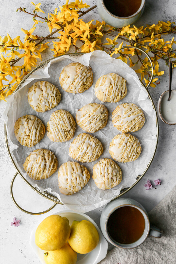 a gold wire basket lined in parchment paper of gluten free iced lemon cookies next to some forsythia blooms, mugs of tea and a bowl of lemons.