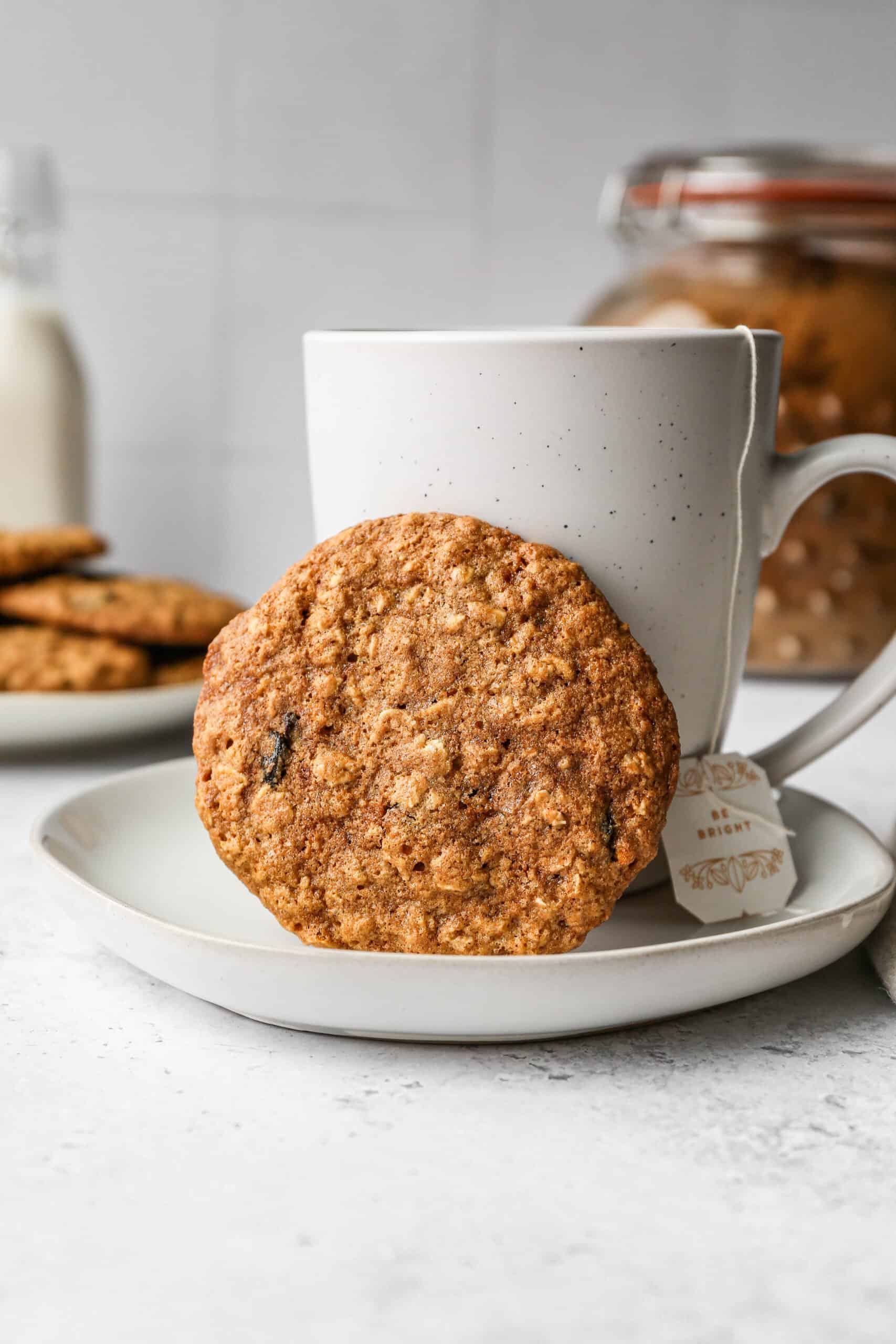 a gluten free oatmeal raisin cookie leaning again a white mug of tea on a white plate.